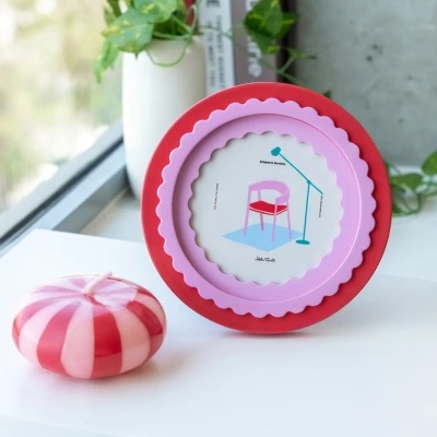 Suporte de cera vermelho e rosa com design de cadeira e candeeiro ao lado de uma vela redonda às riscas vermelhas e brancas, junto a janela.