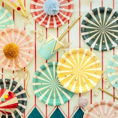 Pratos e copos de festa com riscas e estrelas douradas, chapéus de festa coloridos, e decoração de festa sobre um pano com riscas vermelhas e estrelas douradas.