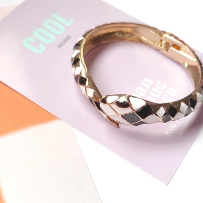 Pulseira dourada com padrão xadrez preto e branco sobre cartão lilás com texto e fundo laranja.