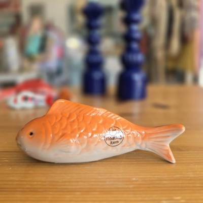 Peixe decorativo de cerâmica laranja e branco em madeira com dois objetos azuis ao fundo.