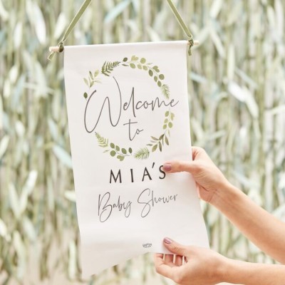 Pendurante de tecido com texto 'Welcome to MIA'S Baby Shower' e moldura de folhas verdes.