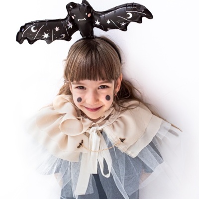 Menina vestida para festa com morcego preto na cabeça e capa bege com tule branco