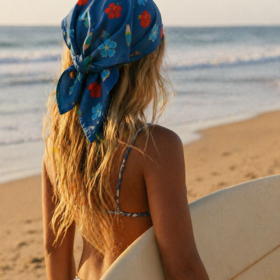 Mulher na praia de costas com lenço azul florido e prancha de surf branca