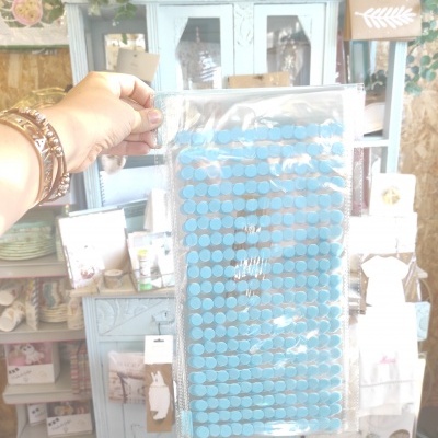 Tapete azul de discos circulares embalado em plástico, segurado por uma mão feminina com pulseiras, fundo com móveis e artigos decorativos.