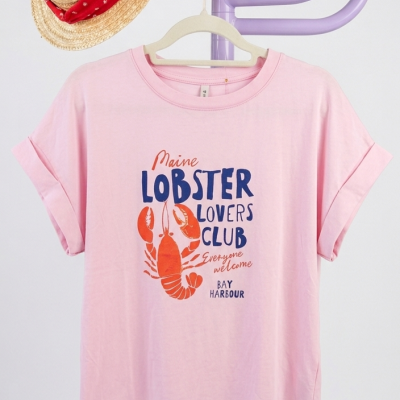 T-shirt rosa com estampa de lagosta e texto, pendurada num cabide creme num suporte lilás com chapéu de palha e lenço vermelho.