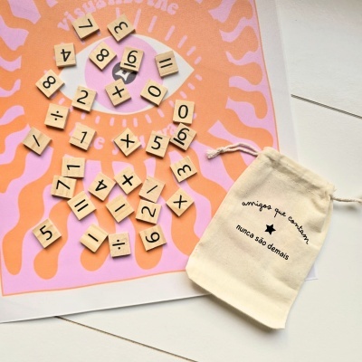 Peças de madeira com números e símbolos matemáticos sobre fundo rosa e laranja e saco de algodão com texto em português