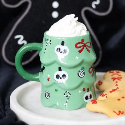 Caneca verde em forma de árvore de Natal decorada com caveiras e doces, com chantilly, em prato com biscoito de gengibre.