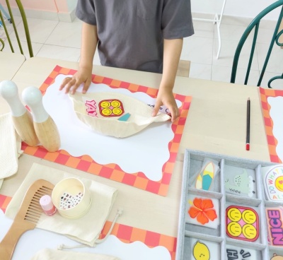 Criança segurando saco de pano com desenho amarelo sorridente e palavra YAY sobre mesa com pentes de madeira, esmalte, adesivos e lápis.