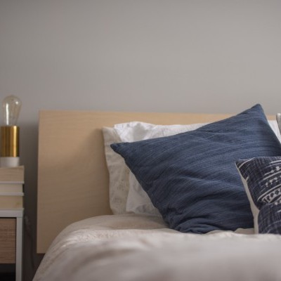 Cama com almofadas azuis e brancas, mesa de cabeceira com luminária e livros, parede cinza