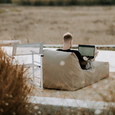 Homem sentado num pufe bege a usar portátil ao lado de cadeira branca em área exterior