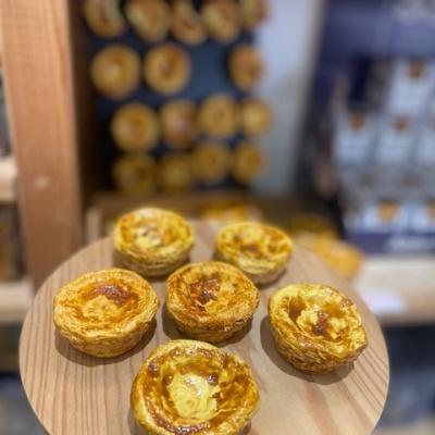 Cinco pastéis de nata num prato de madeira com mais pastéis ao fundo