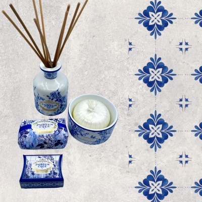 Conjunto de produtos de banho em cerâmica branca e azul com padrão floral e textos decorativos, em fundo de azulejos azuis e brancos.