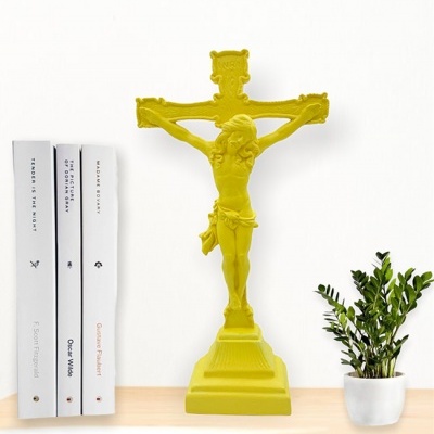 Crucifixo amarelo numa prateleira com livros brancos e planta em vaso branco