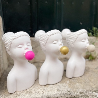 Três esculturas de busto branco com bolas coloridas na boca, em fila sobre pedra