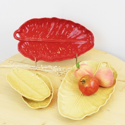 Travessas de cerâmica formato folha vermelho e amarelo com textura e frutos sobre mesa de madeira.