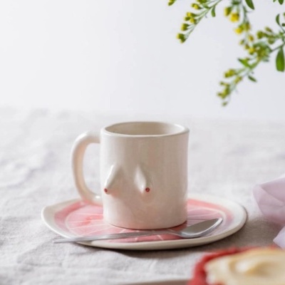 Caneca de cerâmica branca em forma de busto feminino sobre chávena rosa e branca com colher