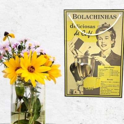 Saco de papel amarelo com texto e imagem, ao lado de jarro com flores num fundo branco