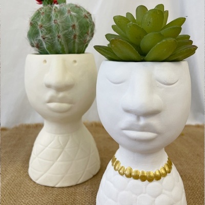Dois vasos de cerâmica em forma de rosto com plantas suculentas, um com um colar dourado e outro com cacto florido