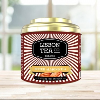 Lata metálica de chá Lisbon Tea Co. bitter almond com tampa dourada e fundo de raios brancos em mesa de madeira