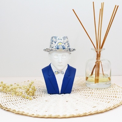 Estátua decorativa de busto humano com chapéu com padrão azul ao lado de frasco de vidro com palitos de difusor