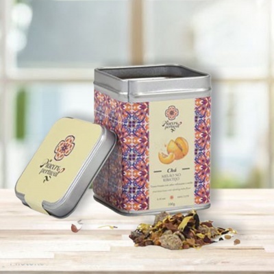 Lata metálica quadrada com chá de melão, laranja e erva-doce, com tampa ao lado e chá solto.