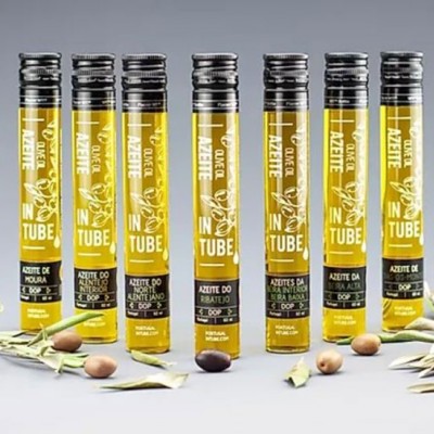 Sete tubos de azeite com rótulos amarelos e tampas pretas em linha, com ramos de oliveira e azeitonas sobre fundo cinzento