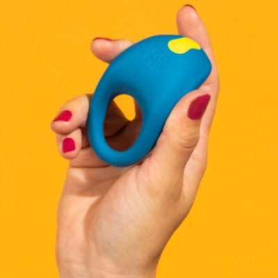 Anel azul de silicone com detalhe amarelo segurado numa mão com unhas vermelhas, fundo amarelo.
