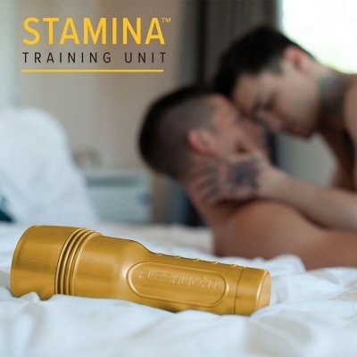 Dispositivo dourado STAMINA TRAINING UNIT sobre cama branca com duas pessoas ao fundo desfocadas.