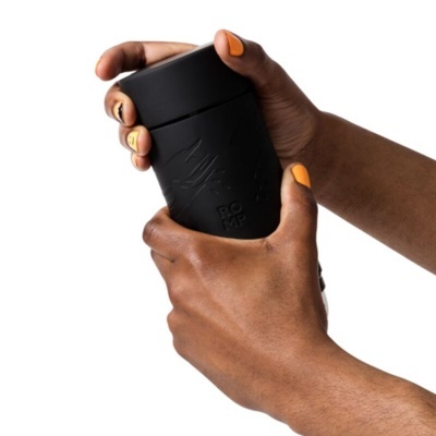 Frasco preto fosco com textura e texto BOMBA segurado por duas mãos com unhas laranja