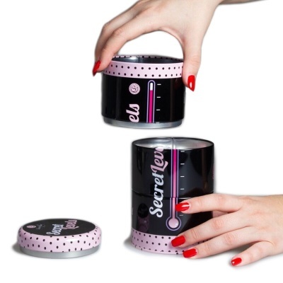 Caixa cilíndrica preta e rosa com poás e texto 'Secrets', mãos com unhas vermelhas seguran a caixa.
