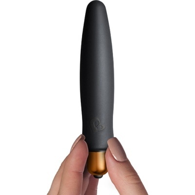 Vibrador pequeno preto com acabamento dourado na base, segurado por mãos em fundo branco