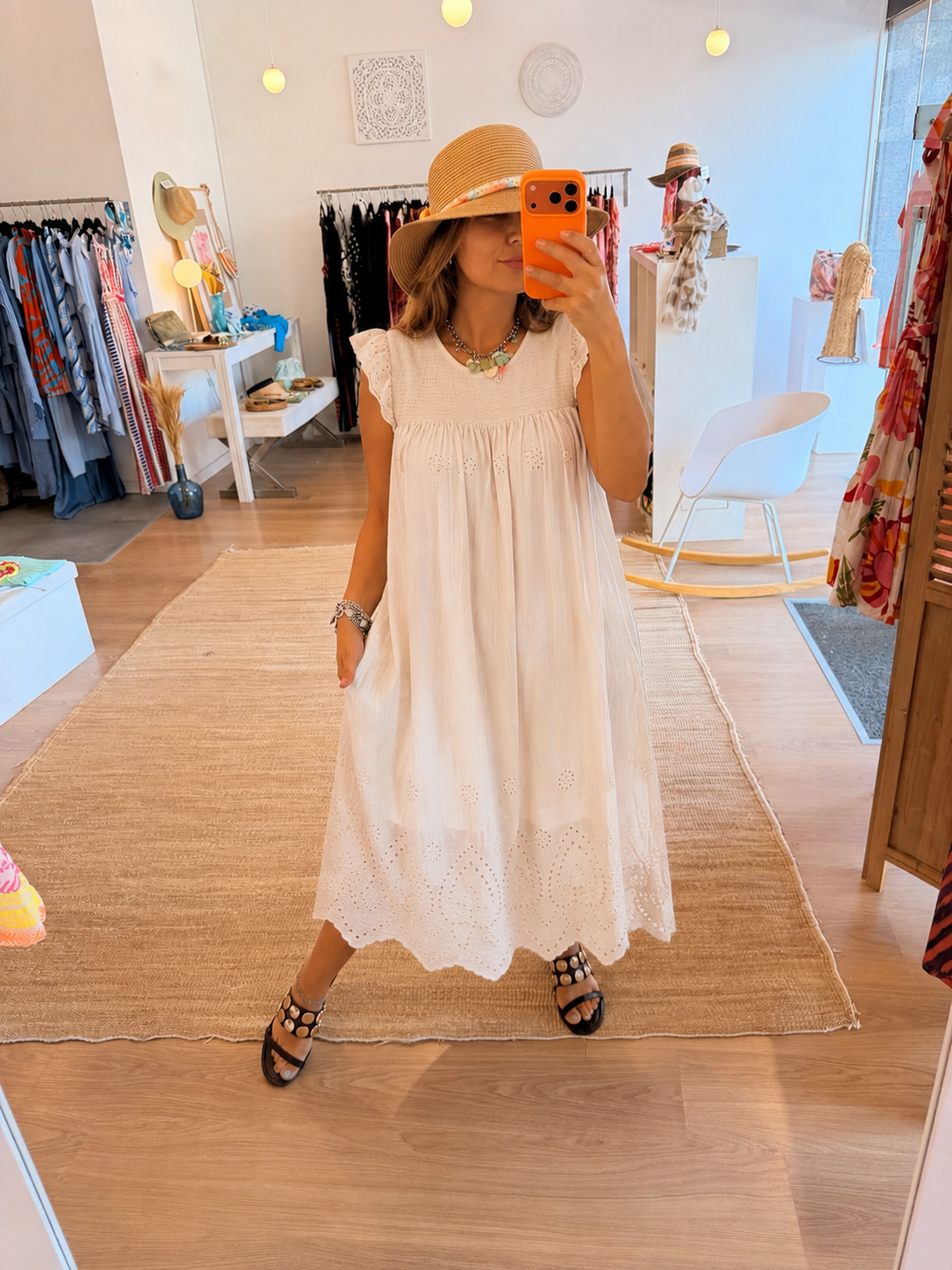 Vestido Encanto Mulher a tirar selfie com vestido branco bordado e chapéu de palha numa loja de roupas