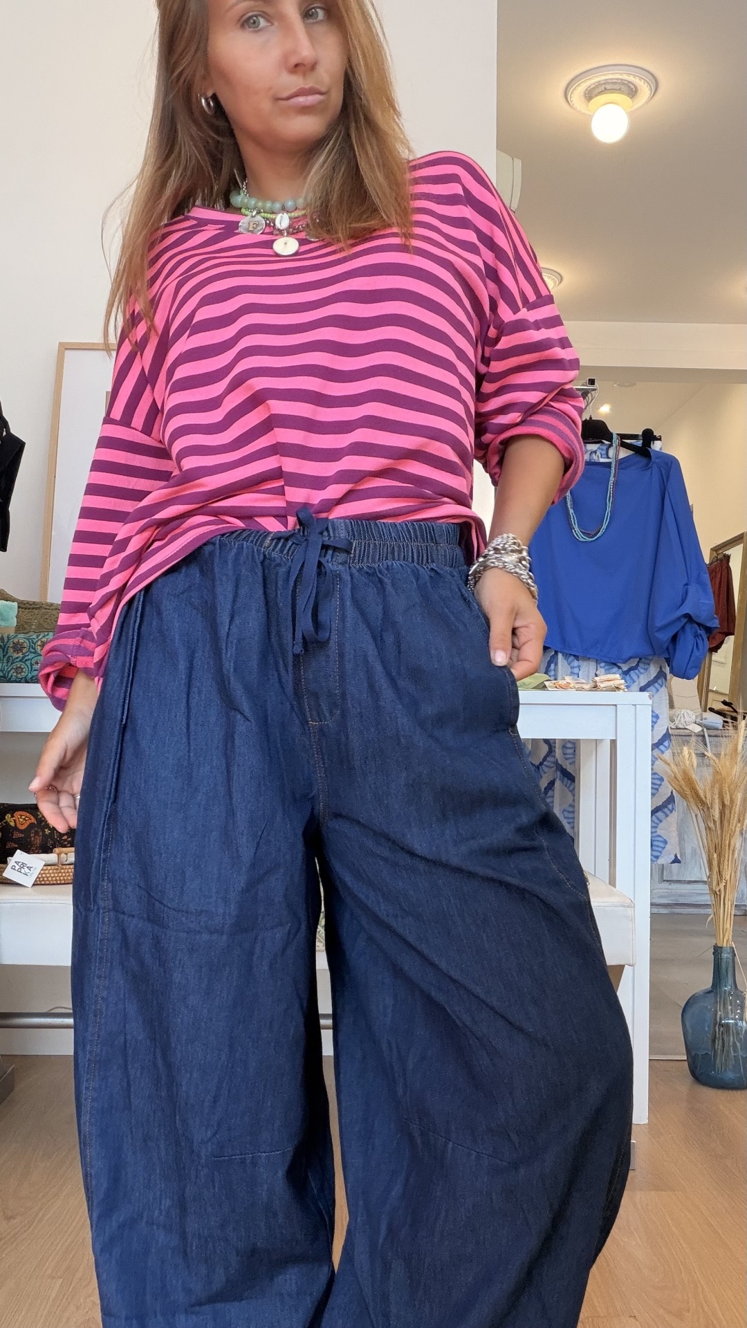 Camisola Riscas Modelo com calças largas de ganga azul escuro e camisa rosa às riscas castanhas, acessórios prateados e colares coloridos, em interior de loja.