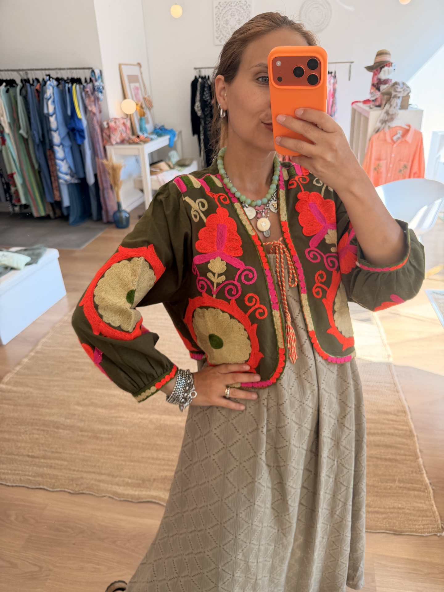 Bolero Bordado Boho Casaco verde-oliva bordado com padrões florais vermelhos e rosa, visto em selfie numa loja de roupa