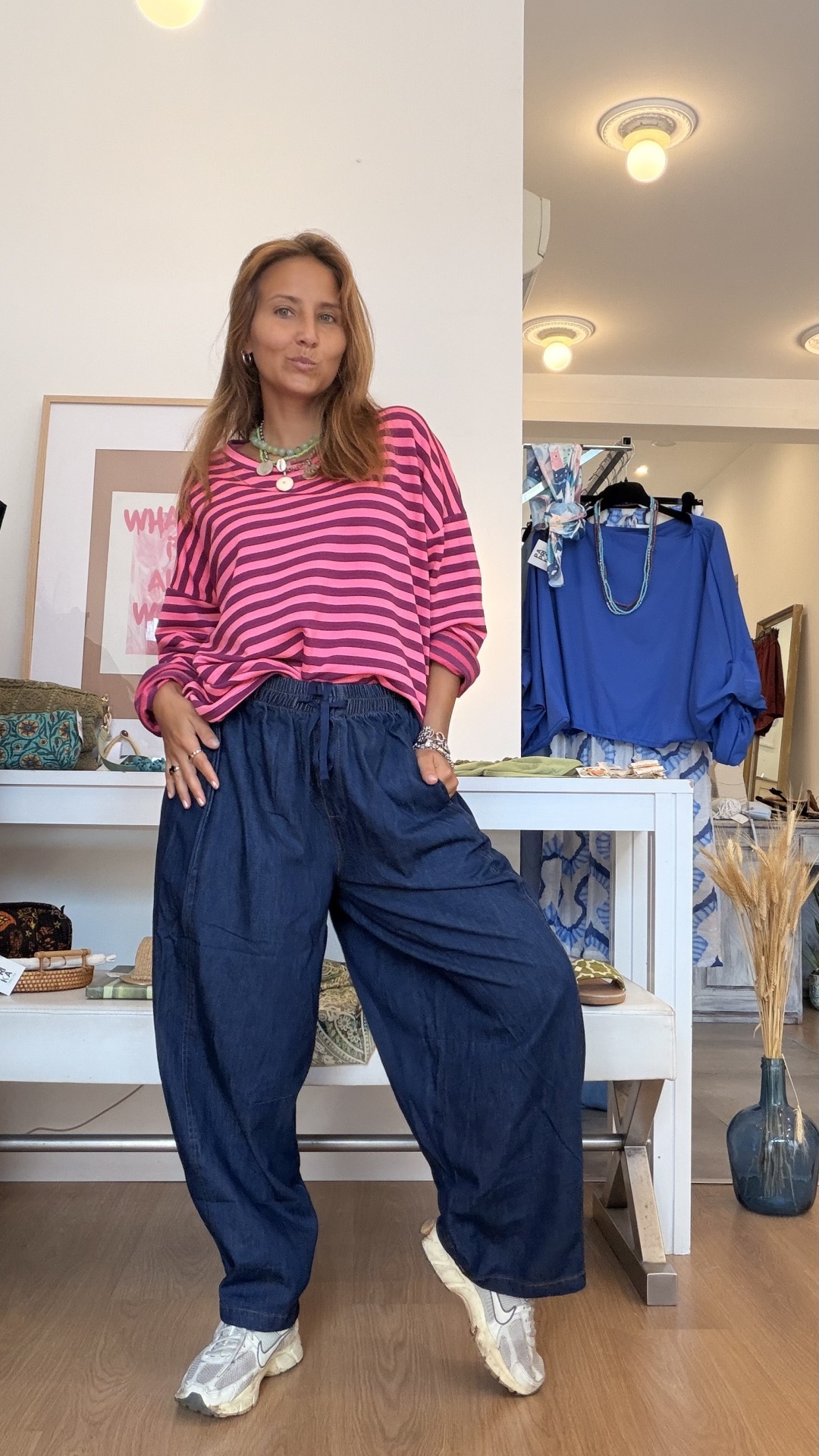 Camisola Riscas Mulher com camisola riscada rosa e preta, calças largas escuras e sapatilhas brancas numa loja de roupas