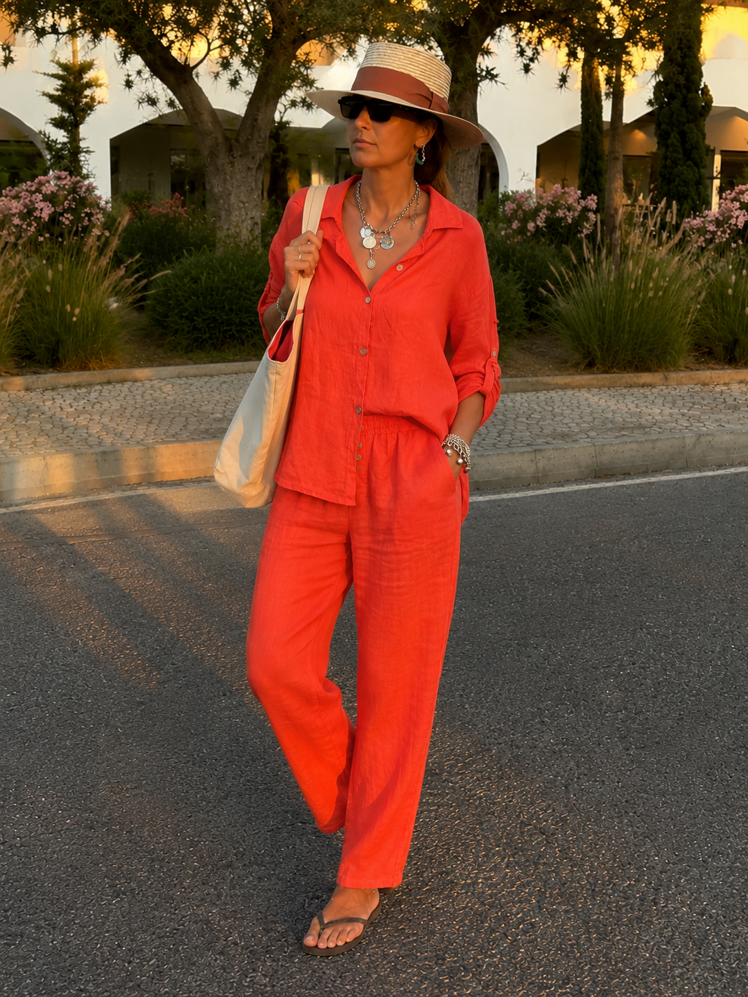 Conjunto Linho Summer Mulher usa conjunto laranja com chapéu de palha, óculos escuros e colares prateados numa rua com vegetação ao fundo.