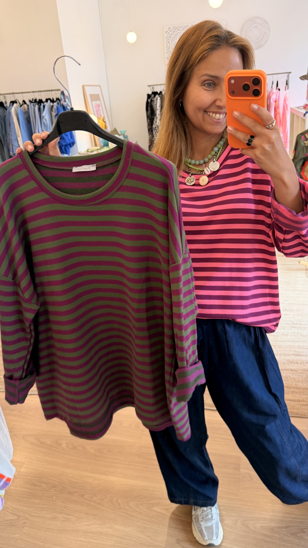 Camisola Riscas Camisola às riscas verde e bordeaux num cabide e mulher a usar camisola às riscas rosa, num ambiente de loja de roupa