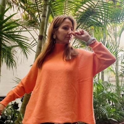 Mulher com camisola laranja e calças de bolas brancas em ambiente com plantas tropicais.