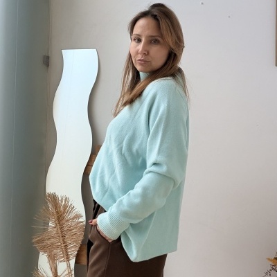 Mulher com camisola azul clara e calças castanhas num ambiente interior minimalista com espelho ondulado