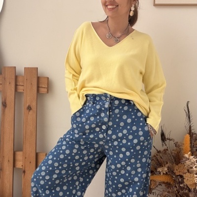 Mulher vestindo camisola amarela e calças azuis com bolinhas brancas, em ambiente interior simples.