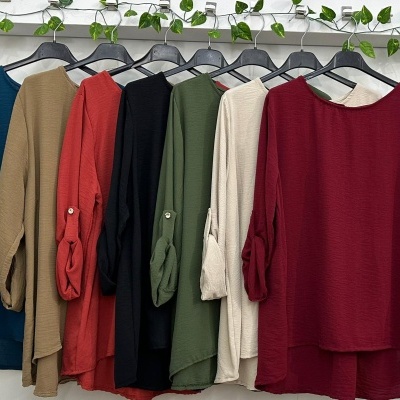 Seis blusas texturizadas em várias cores penduradas em cabides pretos contra parede branca com folhagens.