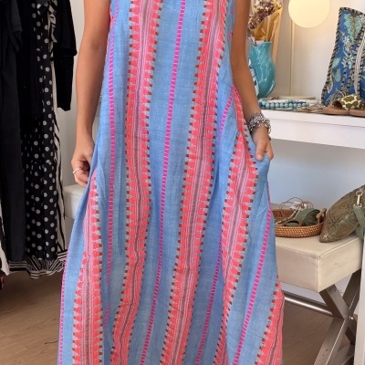Vestido comprido azul com riscas rosas e vermelhas usado por mulher, sandálias brancas e colares, interior com parede branca e mobília.