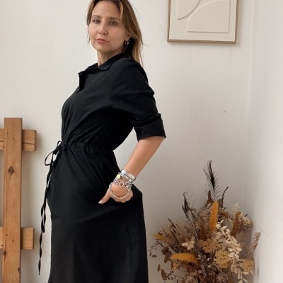 Mulher com vestido preto e botas pretas em ambiente decorado com quadro abstrato e vaso de plantas secas