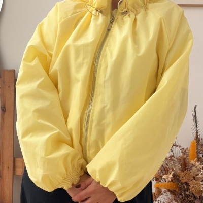 Mulher a usar casaco amarelo com detalhes na gola e punhos, brincos pendentes, fundo com decoração de flores secas