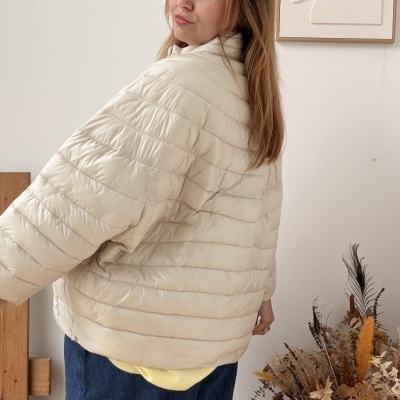 Mulher de casaco acolchoado bege claro e calças azuis largas em ambiente interior com quadro e vaso de flores secas