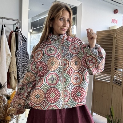 Mulher com casaco estampado colorido e saia vinho em loja com espelho, roupas penduradas e plantas.