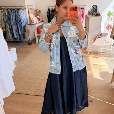 Mulher a tirar selfie num espelho vestindo casaco de ganga azul claro com flores, vestido azul escuro e sandálias claras, dentro de loja de roupa.