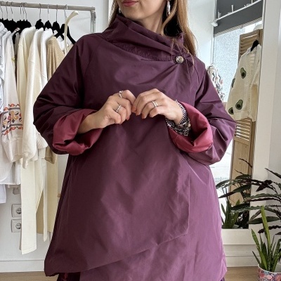 Mulher com túnica ou casaco roxo de tecido impermeável e saia preta com padrão floral vermelho num interior com cabide de roupas, espelho e plantas.