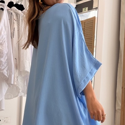 Blusa azul clara larga com mangas 3/4 usada por mulher com jeans e óculos, ambiente interior com cabide e espelho ao fundo.