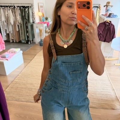Mulher a usar macacão de ganga azul claro com alças castanhas em pele sintética e top castanho, a tirar selfie com telemóvel de capa laranja numa loja de roupa.